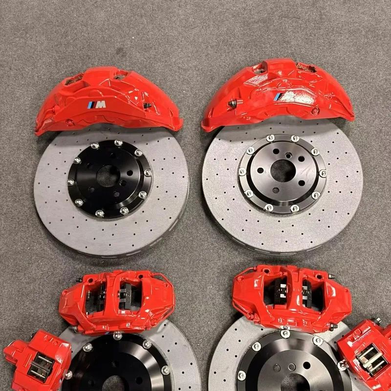 Two red M-marked brake calipers with two drilled rotors with black centre hats and two smaller caliper bodies laid out on a carpet
